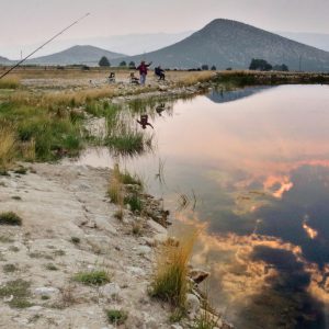 Man fishing on the shores of a pond, mountains in the background. Gentle sunset reflection on the pond