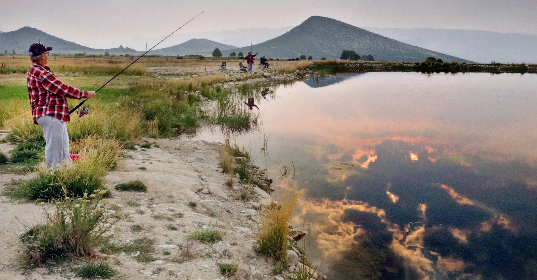 Man fishing on the shores of a pond, mountains in the background. Gentle sunset reflection on the pond