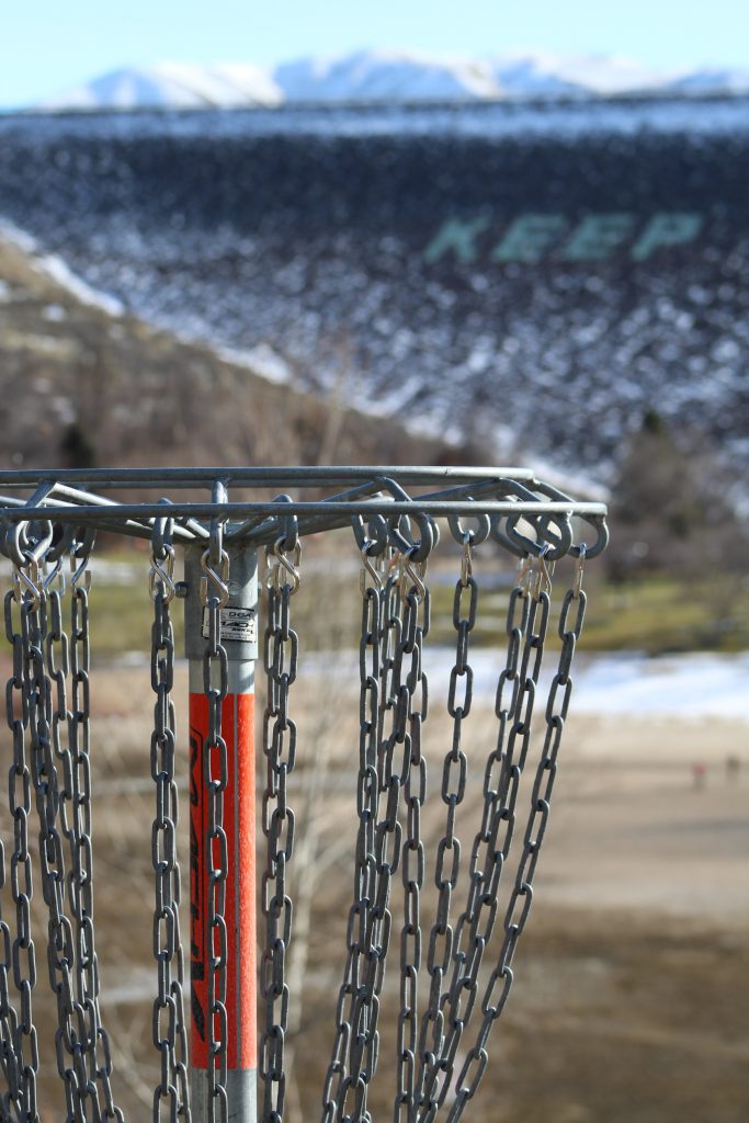 Disc golf basket in foreground, Lucky Peak dam with snowcapped mountains in the background